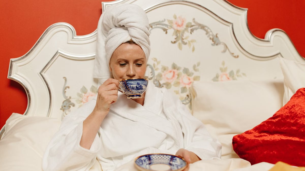 A woman in a white robe and towel wrap enjoys tea in a cozy bedroom setting.